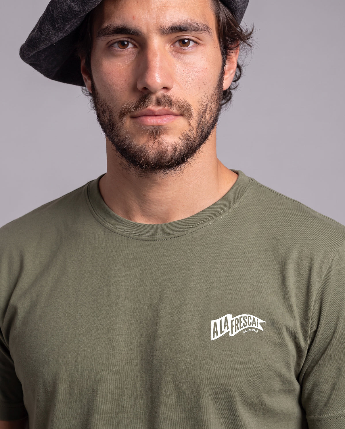 Homem de boina vestindo a Camiseta A La Fresca da Série Sem Alarde em algodão peruano, modelagem masculina, na cor verde, com a bandeirola branca pequena no peito