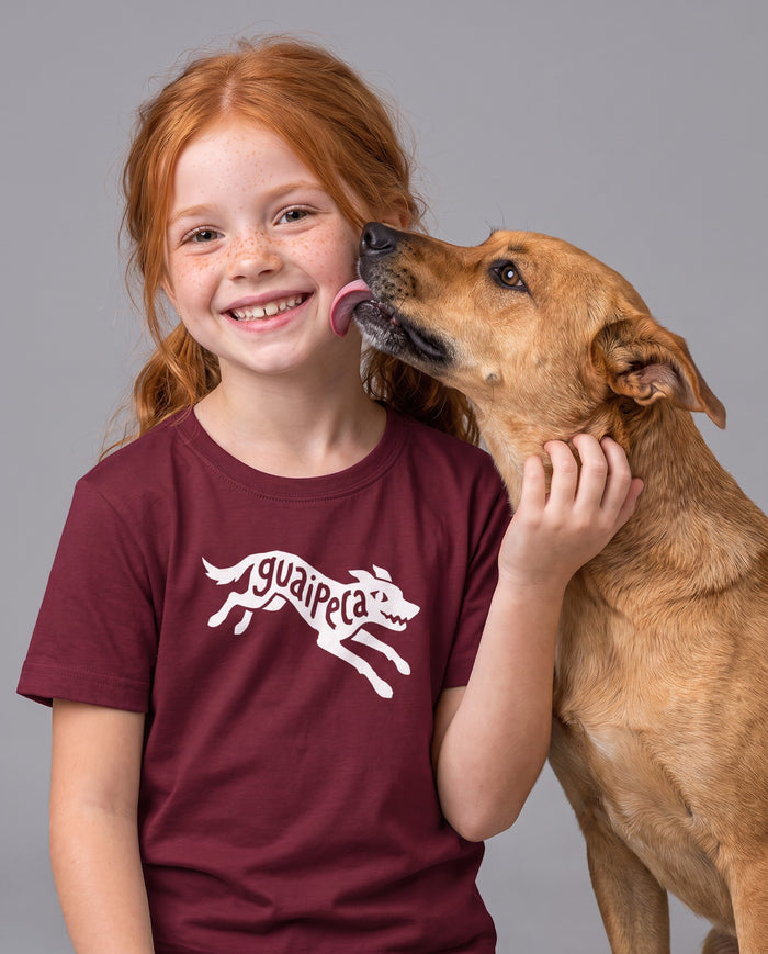 Menina ruiva sorrindo enquanto um cachorro lambe o rosto dela, vestindo a camiseta infantil Guaipeca na cor bordeaux