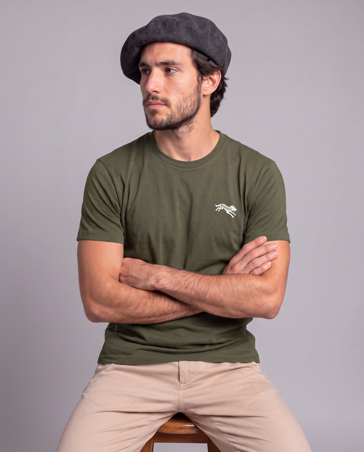 Homem de boina sentado de braços cruzados vestindo a Camiseta Guaipeca da Série Sem Alarde em algodão peruano, modelagem masculina, na cor verde musgo