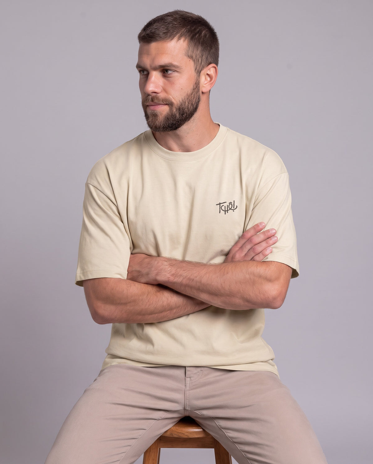 Homem sentado de braços cruzados vestindo a Camiseta Tchê da Série Sem Alarde, modelagem oversized unissex, na cor areia