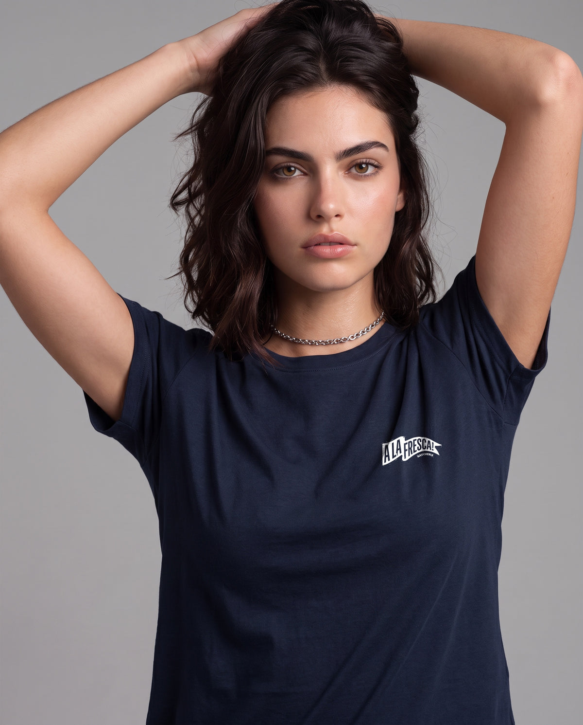 Mulher com as mãos no cabelo vestindo a Camiseta A La Fresca da Série Sem Alarde, modelagem feminina, na cor marinho, com a bandeirola branca pequena no peito