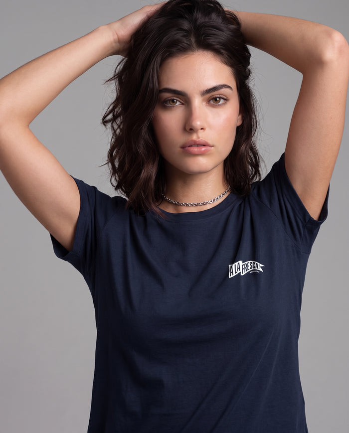 Mulher com as mãos no cabelo vestindo a Camiseta A La Fresca da Série Sem Alarde, modelagem feminina, na cor marinho, com a bandeirola branca pequena no peito