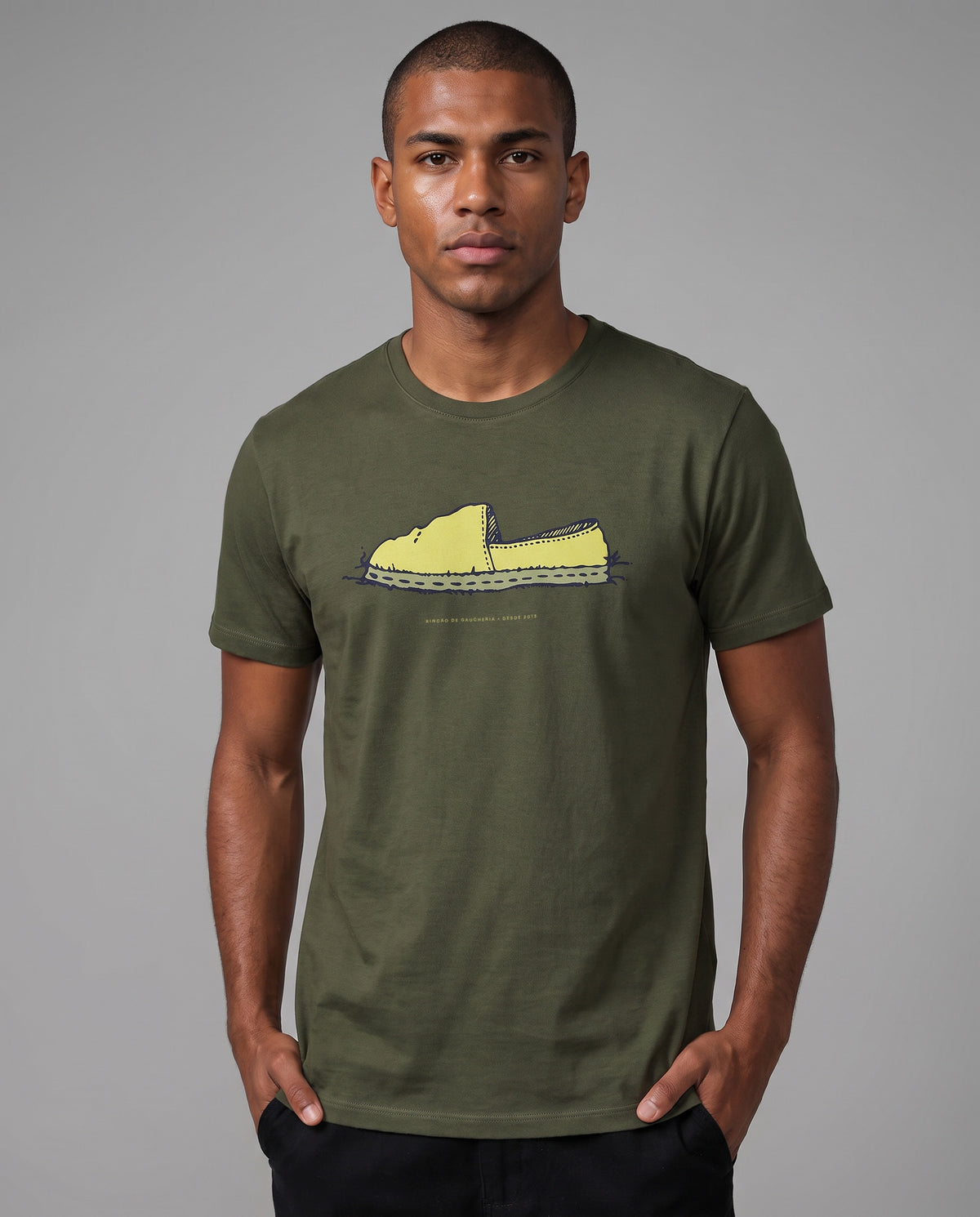 Homem com as mãos no bolso vestindo a Camiseta Alpargata Identidade de Campo em algodão peruano, modelagem masculina, na cor verde, com a estampa da alpargata no peito