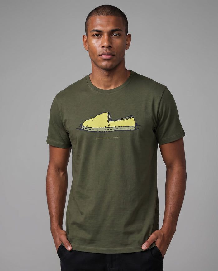 Homem com as mãos no bolso vestindo a Camiseta Alpargata Identidade de Campo em algodão peruano, modelagem masculina, na cor verde, com a estampa da alpargata no peito