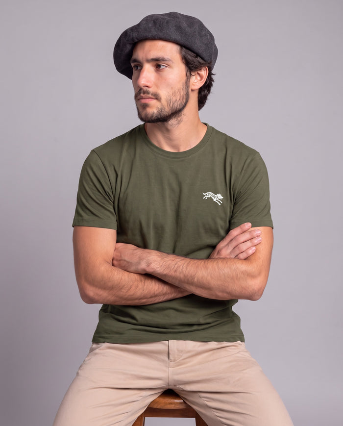 Homem de boina sentado de braços cruzados vestindo a Camiseta Guaipeca da Série Sem Alarde em algodão peruano, modelagem masculina, na cor verde musgo
