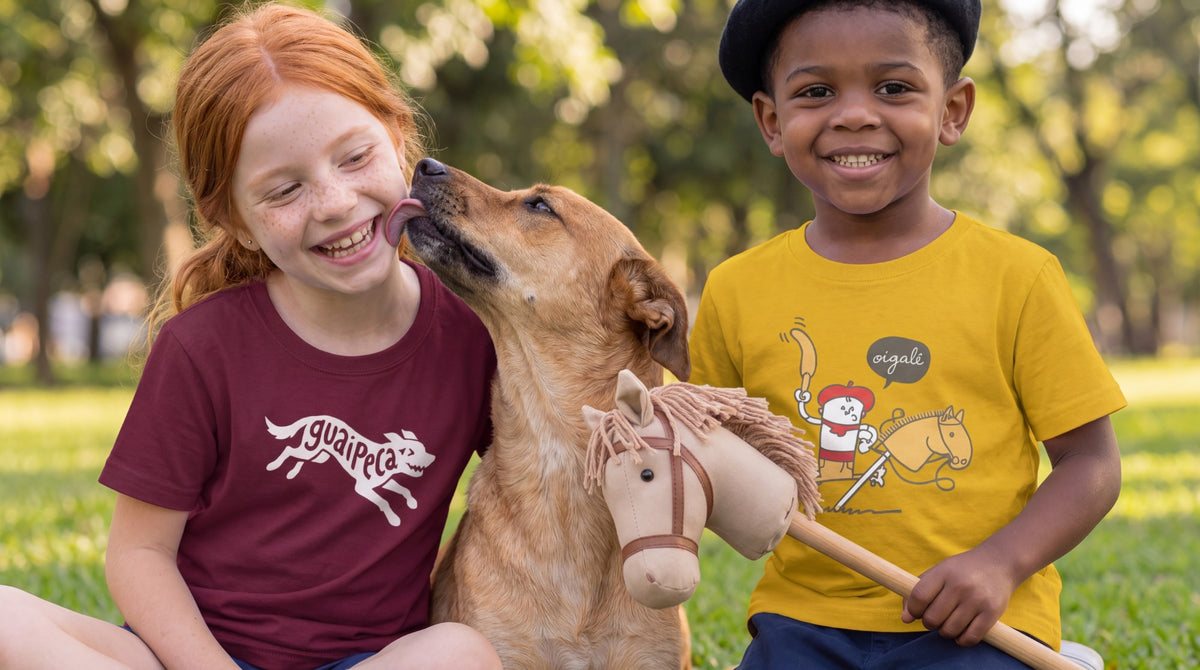 Duas crianças com camisetas infantis da Gaucheria, Guaipeca e Oigalê, rindo com um cachorro na grama