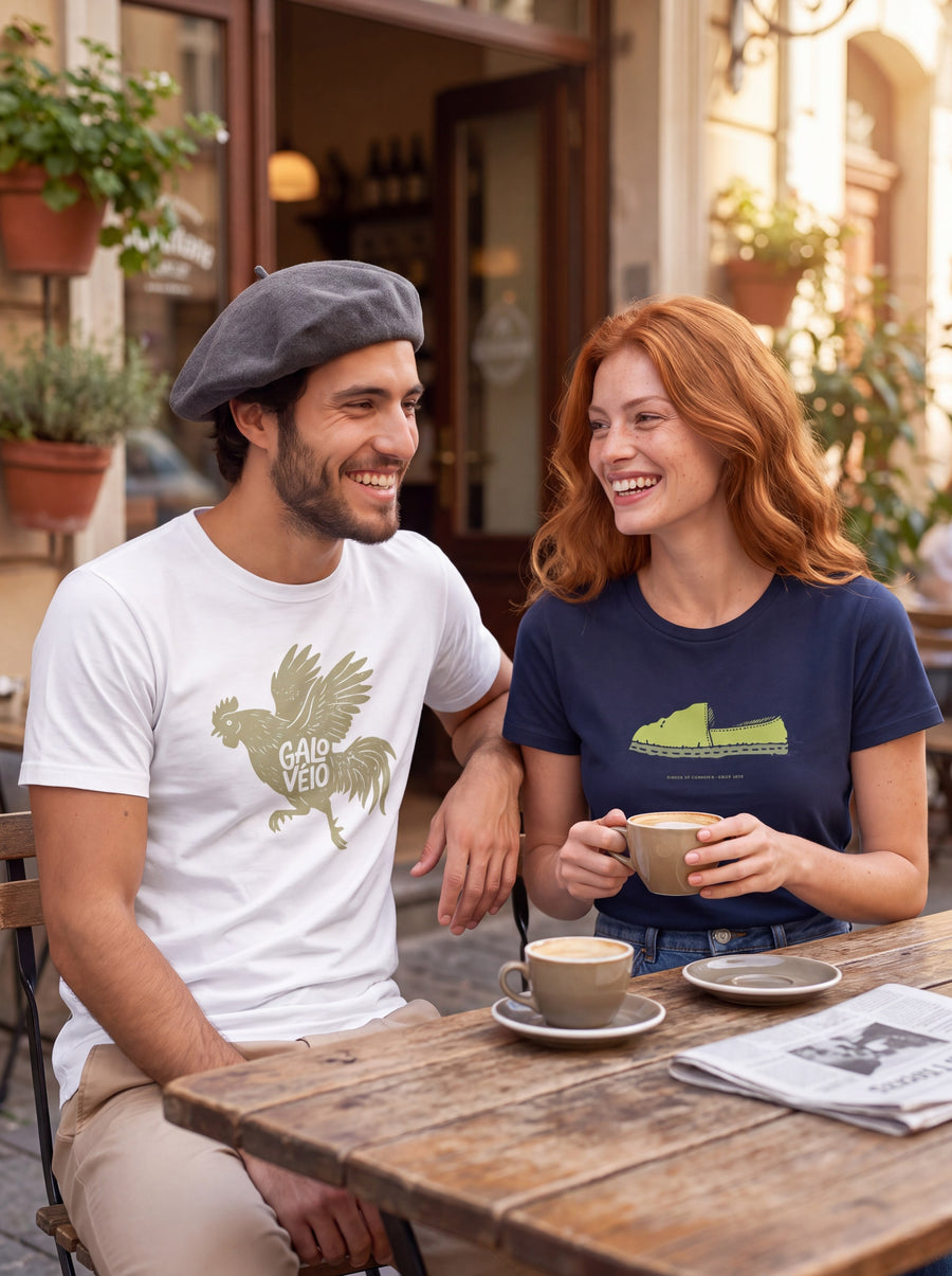 Casal rindo na mesa de um café, ele de boina com a camiseta Galo Véio branca e ela com a camiseta Alpargata azul marinho